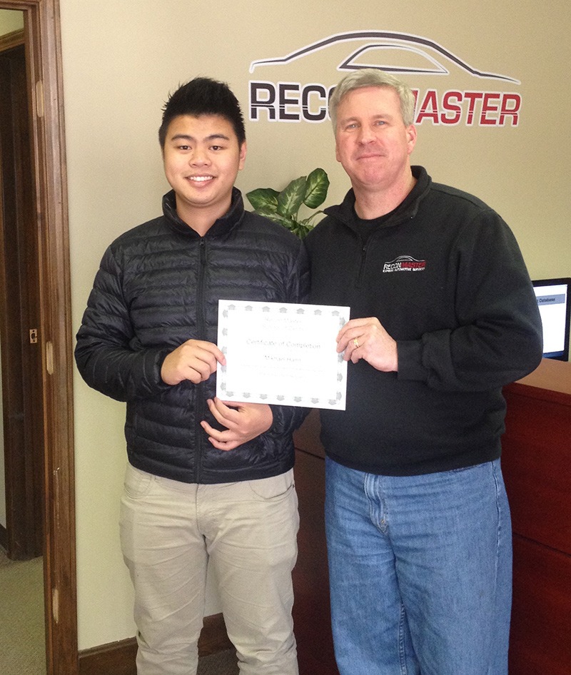 A young man and an older man pose together in an office, proudly holding a certificate of completion. The setting indicates a professional achievement.