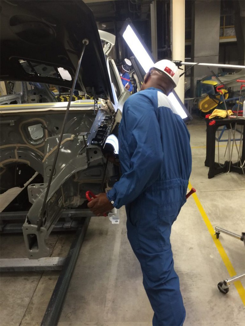A technician in a blue jumpsuit works on a vehicle's frame in a workshop, using tools to adjust the structure under bright overhead lights.