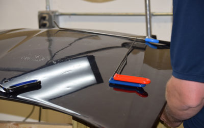 A person secures automotive bodywork using clamps on a glossy black car hood, highlighting maintenance or restoration efforts in a workshop.