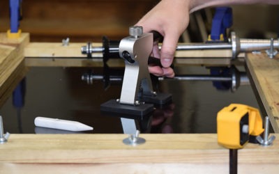 A hand operates a tool on a glossy black surface within a wooden frame, likely part of a DIY project or workshop setup.