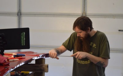 A man with a beard works focusedly on a large red object in a garage, using tools to refine or repair it. The scene highlights craftsmanship.
