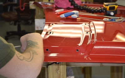 A person with a tattoo works on a shiny red car part, surrounded by tools, highlighting vehicle restoration in a workshop setting.