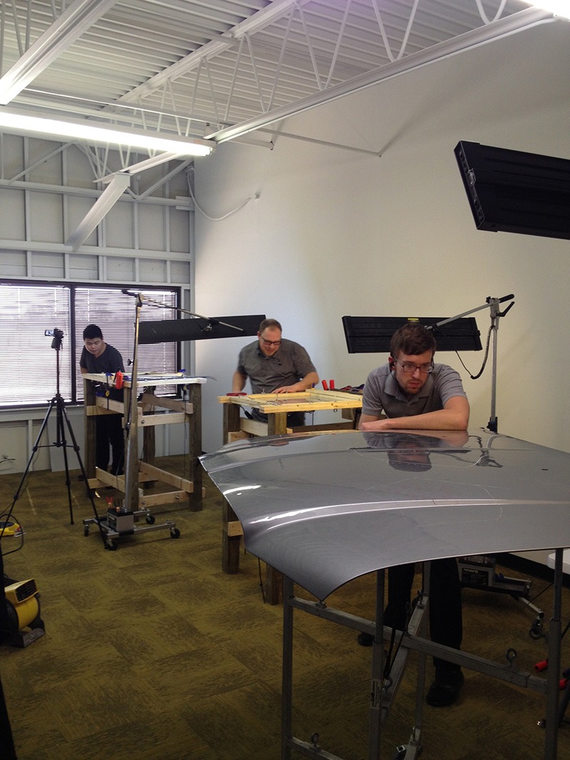 Three technicians work in a well-lit workshop, focusing on a gray automotive hood displayed on a stand, surrounded by equipment and tools.