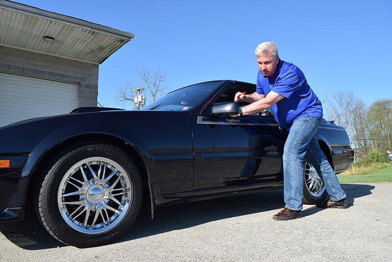 A man in a blue shirt leans against a black turbocharged car, reaching to open the driver's door in a sunny, outdoor setting.