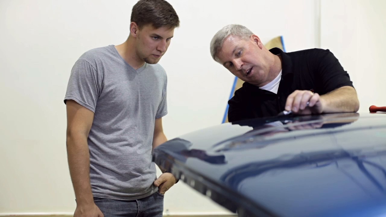 A man instructs a young man on car maintenance, focusing on the vehicle's hood in a workshop setting. The scene emphasizes hands-on learning.