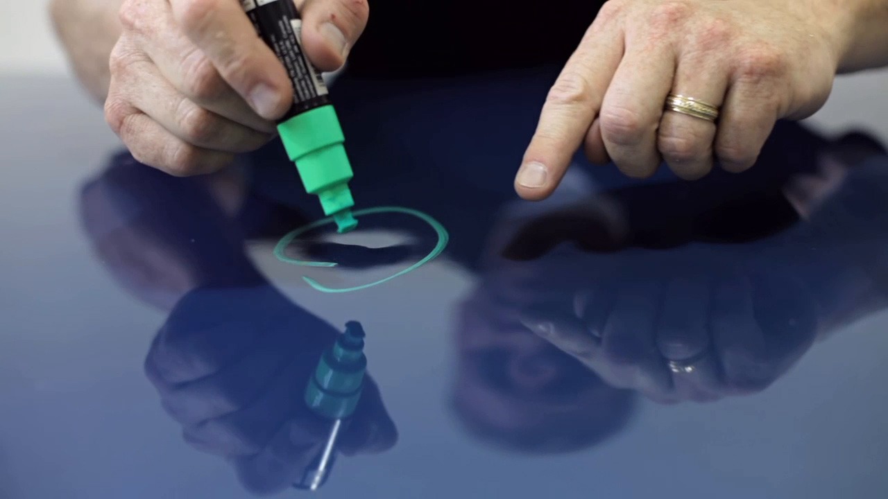 A hand holds a green marker, drawing a circle on a glossy surface while reflecting light. This illustrates surface marking techniques.